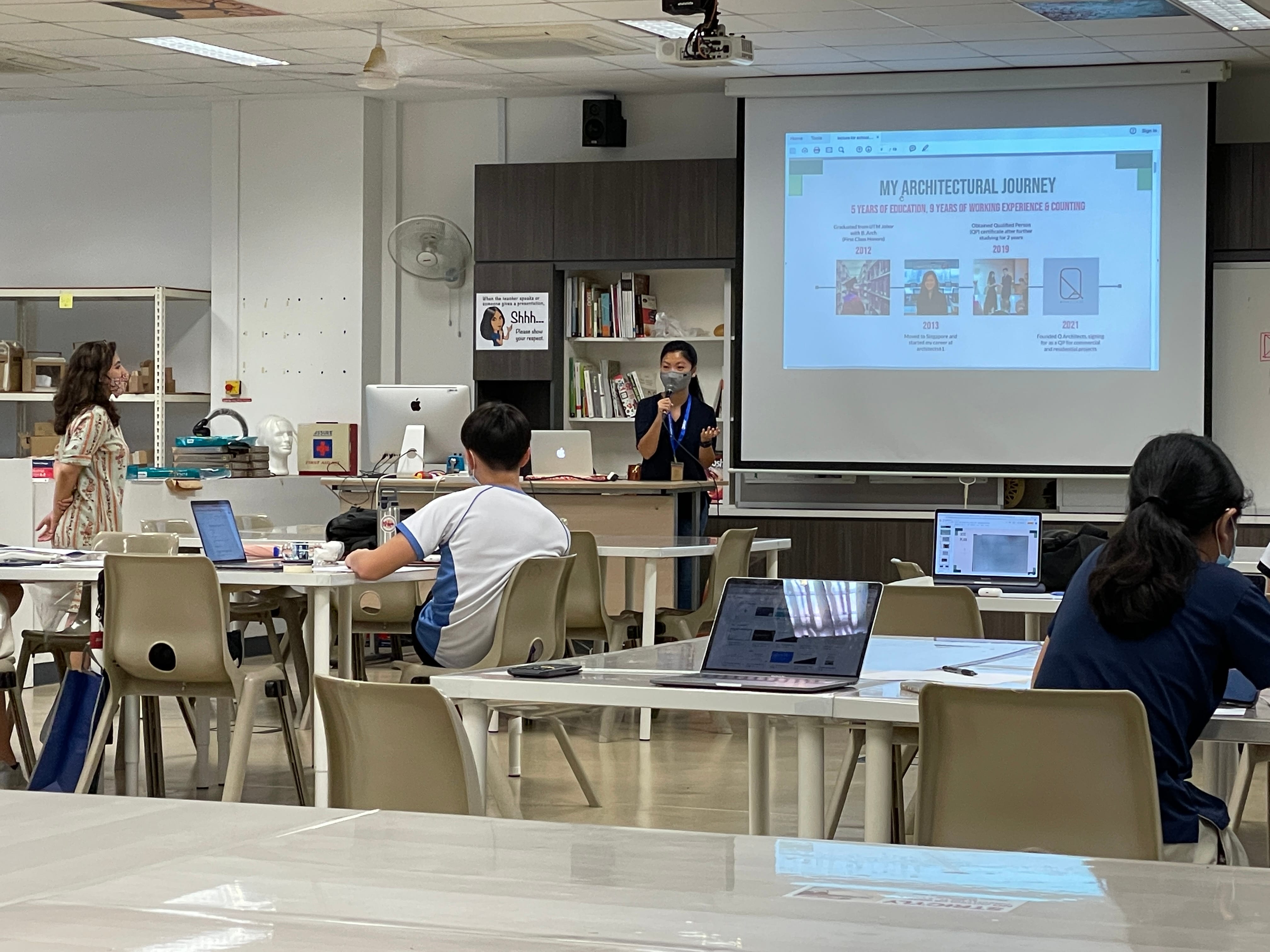 A masked presenter speaks in a classroom showing a projected slide titled "My Architectural Journey" to seated students with laptops.